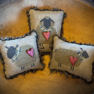 May include: Three decorative sheep pillows with cross-stitch details. Each pillow features a sheep design with a heart accent. The pillows have a neutral color palette with a frayed edge, and are arranged in a wooden bowl.