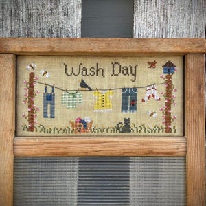 Peut inclure: Une broderie au point de croix encadrée en bois avec un fond beige et le texte "Wash Day". La broderie représente un fil à linge avec divers vêtements suspendus, dont une salopette, une chemise, une robe et un pantalon. On y voit également deux oiseaux, un chat et une petite maison.