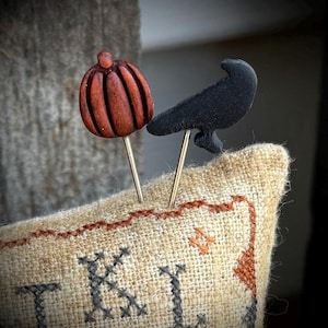 May include: Two decorative pins featuring a carved brown pumpkin and a solid black raven silhouette. The pins are inserted into a piece of tan burlap fabric with visible cross-stitch embroidery in black and orange thread along the top edge.