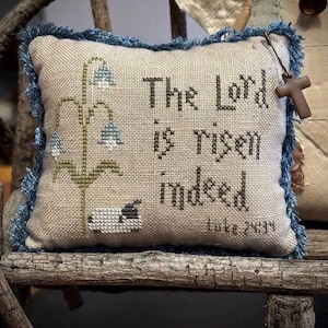 Può includere: Cuscino quadrato decorativo con la frase ricamata "The Lord is risen indeed". Presenta una frangia blu e una piccola croce di legno. Il design include un motivo floreale e una pecora. Il cuscino è in tessuto beige.