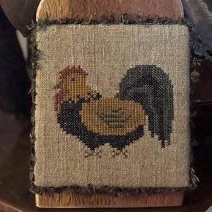 Puede incluir: Un patrón de punto de cruz de un gallo marrón, negro y rojo sobre un fondo de lino beige. El gallo está bordado con una cola negra, un cuerpo marrón y una cresta y barbillas rojas.
