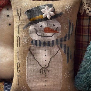 May include: A rectangular tan fabric cushion featuring a cross-stitched snowman wearing a dark grey top hat with a white snowflake button, a striped scarf, and an orange nose. The word WINTER is stitched vertically in black on the left side.