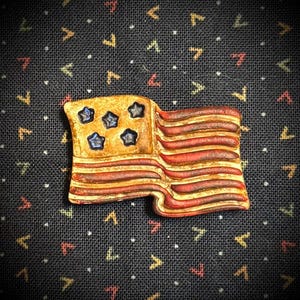 May include: A handcrafted pin shaped like the American flag. The pin features a gold-colored rectangle with five blue stars and red and brown stripes. The pin is set against a dark blue fabric background with a repeating pattern.