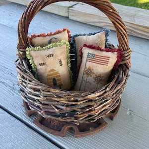 Peut inclure: Un panier en osier avec une poignée brune rempli de quatre petits coussins brodés. Les coussins ont des motifs différents, notamment des abeilles, une maison, des tournesols et un drapeau américain.