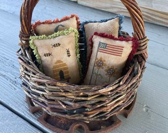 May include: A wicker basket with a brown handle filled with four small embroidered pillows. The pillows have different designs, including bees, a house, sunflowers, and an American flag.