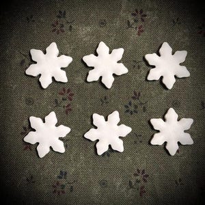 May include: Six white snowflake-shaped ornaments arranged on a dark green fabric with a floral pattern. The snowflakes have six points and a slightly textured surface, perfect for winter crafts or decorations.