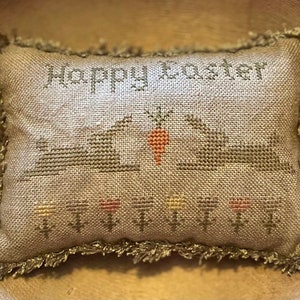 May include: A beige pillow with a frayed edge, embroidered with the text "Happy Easter" and a scene of three bunnies with a carrot.