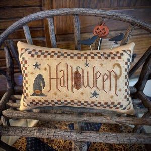 May include: A rectangular beige cross-stitch cushion featuring the word Halloween in brown lettering, a witch hat, stars, and a checkered border. Three small decorative picks shaped like a pumpkin, a bat, and a bird are inserted into the top.