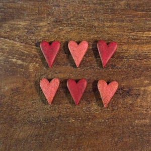 May include: Six small, heart-shaped wooden ornaments in varying shades of red are arranged on a rustic, brown wooden surface. The hearts have a slightly textured appearance, suggesting a handmade or vintage quality. The arrangement is symmetrical.