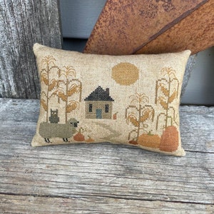 Puede incluir: Una almohada beige con una escena de otoño bordada. La escena incluye una pequeña casa, un gato negro, una oveja, tallos de maíz, calabazas y un sol.