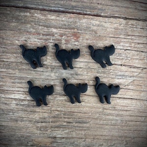 May include: Six small, black cat-shaped embellishments are arranged on a weathered, wooden surface. Each cat has a simple, stylized design with a curved tail and pointed ears. The cats are evenly spaced, creating a pattern.