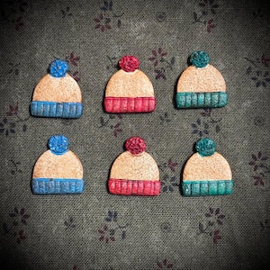May include: Six miniature winter hat ornaments. Each hat has a tan crown, a ribbed brim, and a pom-pom. The brims are blue, red, and teal, and the pom-poms match the brim colors. The ornaments are arranged on a patterned green background.