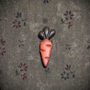 May include: A small, orange carrot-shaped object with a dark brown top. The carrot has carved lines to represent the texture. The background is a textured, olive-green fabric with small floral patterns in shades of purple and blue.