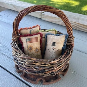 May include: A wicker basket with a brown handle sits on a wooden surface. Inside the basket are four small, square, fabric pillows with embroidered designs. The designs include a crescent moon, a snowman, a sunflower, and an American flag.