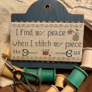 May include: A blue wooden sign with a cross-stitch design that reads "I find my peace when I stitch my piece". The sign is surrounded by wooden spools of thread, some painted green, and a pair of small scissors.