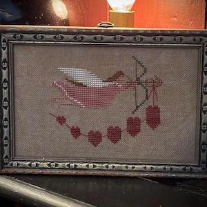 Può includere: Opera d'arte a punto croce incorniciata raffigurante un Cupido rosa con arco e frecce, circondato da una ghirlanda di cuori rossi. L'opera è ambientata su uno sfondo beige e incorniciata in una cornice di legno decorativa.