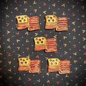 May include: Five small, handcrafted flag-shaped decorative items. Each flag features a yellow field with blue dots and red and white stripes, resembling the American flag. The flags are arranged on a dark patterned background.