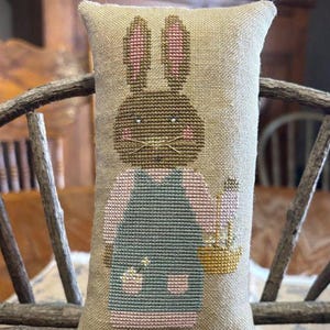 May include: A rectangular, cross-stitched pillow featuring a brown bunny wearing a blue and pink outfit and holding a basket. The bunny has pink ears and cheeks. The pillow is beige and rests on a wooden bench.