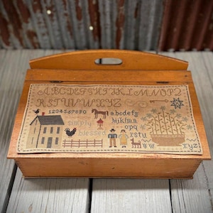 Peut inclure: Une boîte en bois avec un couvercle à charnières et un motif de point de croix sur le devant. Le motif représente une maison, un cheval, une poule et l'alphabet. Le texte "Simply Blessed" est également inclus dans le motif.
