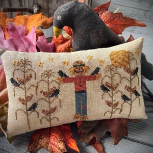 May include: A rectangular, beige cross-stitch pillow featuring a scarecrow, corn stalks, and black birds. The scarecrow wears a red shirt and blue pants. A black crow figurine sits behind the pillow, surrounded by fall leaves.