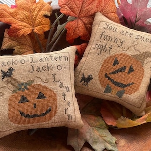 Pode incluir: Duas almofadas pequenas, quadradas, de estopa com rostos de abóbora talhada bordados. Uma almofada diz "Jack-o'-Lantern Jack-o-Lantern" e a outra diz "You are such a funny sight". As almofadas estão em uma tigela de madeira com folhas de outono.