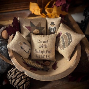 May include: A collection of small, burlap-textured pillows with embroidered designs, including a star, a pilgrim, a turkey, and a pumpkin. One pillow reads "Come with thankful hearts." The pillows are arranged in a wooden bowl with fall foliage.