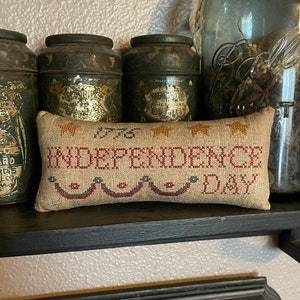 May include: A small, beige pillow with red, white, and blue embroidery. The pillow reads "1776 Independence Day" with stars and a banner design.