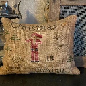 May include: A small, burlap cushion featuring cross-stitch Christmas designs. The design includes a Santa Claus figure, reindeer, Christmas trees, and the words "Christmas is coming..." in brown thread. The cushion has a neutral colour palette.