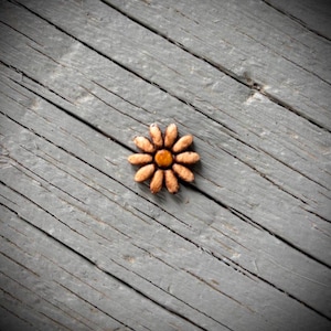 May include: A wooden daisy with a brown center and tan petals rests on a weathered gray wooden surface. The flower is the focal point, with the diagonal wood grain adding texture and contrast.
