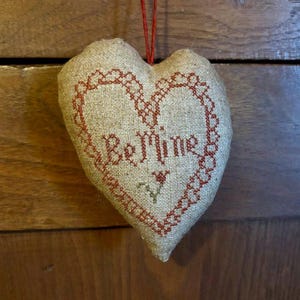 May include: A heart-shaped pillow with a beige linen fabric and red embroidery. The embroidery features a heart with the words "Be Mine" inside.