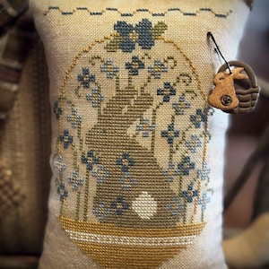 May include: A decorative pillow with a cross-stitch design featuring a bunny in a basket surrounded by blue flowers. The pillow has a beige background with a gold border and a small bunny charm attached. The pillow measures approximately 10 inches in length.