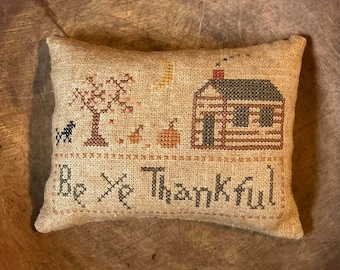 Puede incluir: Una almohada beige con un diseño de punto de cruz que presenta una pequeña cabaña, un árbol y calabazas. El texto "Be Ye Thankful" está bordado debajo del diseño.