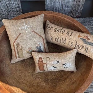 May include: Four embroidered throw pillows in a wooden bowl. The pillows feature a nativity scene, a shepherd with sheep, a Christmas angel, and the text "For unto us a child is born".