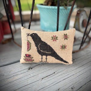 Peut inclure: Un petit coussin rectangulaire beige avec un motif de corbeau noir. Le corbeau tient une tranche de pastèque dans son bec et se tient près de piles de tranches de pastèque. Des accents floraux roses et verts sont brodés sur le coussin.