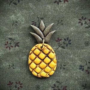 May include: A gold-colored pineapple pendant with a textured surface and a silver-colored crown. The pendant is hanging on a gold-colored loop. The background is a green fabric with a floral pattern.