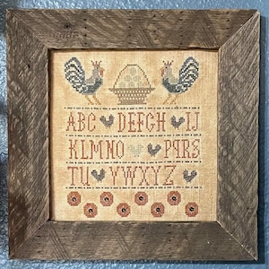 May include: A framed cross-stitch piece with a rustic wooden frame. The cross-stitch features two roosters, a basket of wheat, and the alphabet with hearts between each letter. The letters are in brown and the hearts are in red.