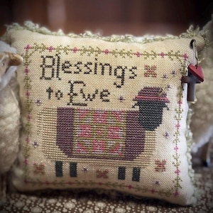 May include: A decorative pillow with the embroidered text "Blessings to Ewe" and a cross-stitched sheep wearing a hat. The sheep is in shades of purple, pink, and green. The pillow has a cream-colored background with a decorative border.