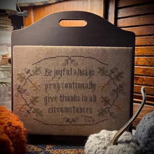 May include: A dark brown wooden carrier featuring a rectangular cross-stitch sampler on a tan fabric background. The text reads: Be joyful always, pray continually, give thanks in all circumstances, 1 Thess 5:16-18, surrounded by stitched leaves.