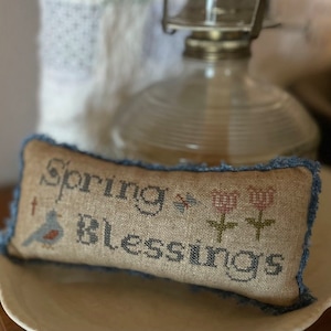 May include: A small, rectangular pillow with a blue denim border. The pillow is made of a beige linen fabric and features embroidered text that reads "Spring Blessings" along with a small bird and two stylized tulips.