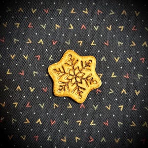May include: A snowflake-shaped button with a detailed snowflake design in the center. The button is a light brown color and sits on a dark blue fabric with a repeating pattern of small white dots and colorful shapes.