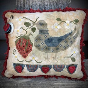 Peut inclure: Un coussin de couleur crème avec une bordure de frange rouge présente un motif de point de croix d'un oiseau bleu avec une fraise dans son bec. L'oiseau est entouré de fleurs blanches et de feuilles vertes. Le bas du coussin est orné d'une rangée de fraises rouges.