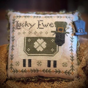 May include: A cross-stitched pillow with the words "Lucky Ewe" and a sheep wearing a green leprechaun hat. The sheep has a green clover design on its back. The pillow has a green and gold trim and a small hat charm.