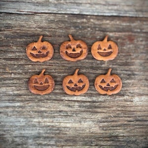 May include: Six small, light brown, flat pumpkin-shaped cutouts arranged in two rows of three. Each pumpkin features a dark, laser-engraved jack-o'-lantern face with triangular eyes and a wide, grinning mouth with a single tooth visible in the center.