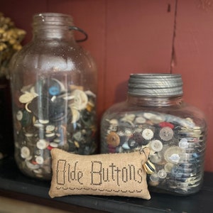 Peut inclure: Deux bocaux en verre remplis de boutons de différentes formes et couleurs, l'un avec un couvercle argenté. Un petit coussin avec le texte "Old Buttons" est devant les bocaux.