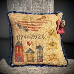 May include: A decorative, square pillow with a tan fabric base and a dark blue trim. The pillow features cross-stitch embroidery of a flag, stars, houses, and the text "1776-2026". A small bell hangs from the top.