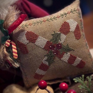 May include: A small, square, tan pillow with a cross-stitched design of two candy canes and a sprig of holly. The candy canes are red and white, and the holly is green. A red ribbon and a candy cane ornament are attached to the pillow.