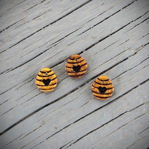 May include: Three decorative, oval-shaped buttons with a bee-hive design. The buttons are yellow and brown with black heart-shaped cutouts. The buttons are arranged on a gray, wooden surface.