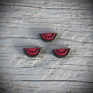 May include: Three watermelon-shaped decorative objects. Each has a red interior with black seed details and a dark gray rind. The objects are arranged on a weathered, gray wooden surface.