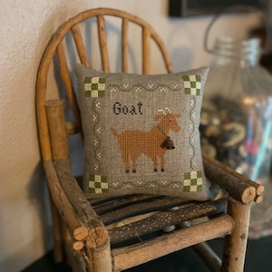Könnte beinhalten: Ein braunes und weißes besticktes Kissen mit einem Ziegenmotiv und dem Wort "Goat" darauf gestickt. Das Kissen liegt auf einem Holzstuhl.