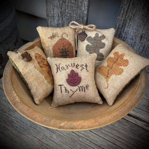 May include: A collection of small, burlap-textured pillows with embroidered autumn-themed designs, including leaves, a pumpkin, and the words "Harvest Thyme." The pillows are arranged in a wooden bowl, creating a rustic, seasonal display.